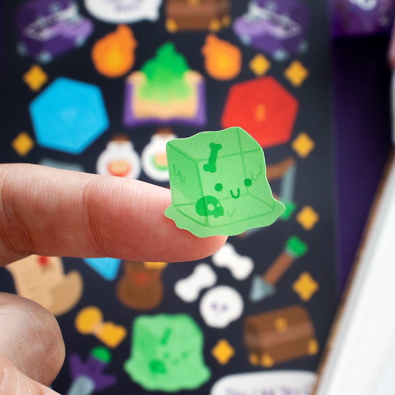 May include: A green slime cube sticker with a skull and bone inside. The sticker has a cute cartoon face with a smile and closed eyes. The sticker is on a finger.