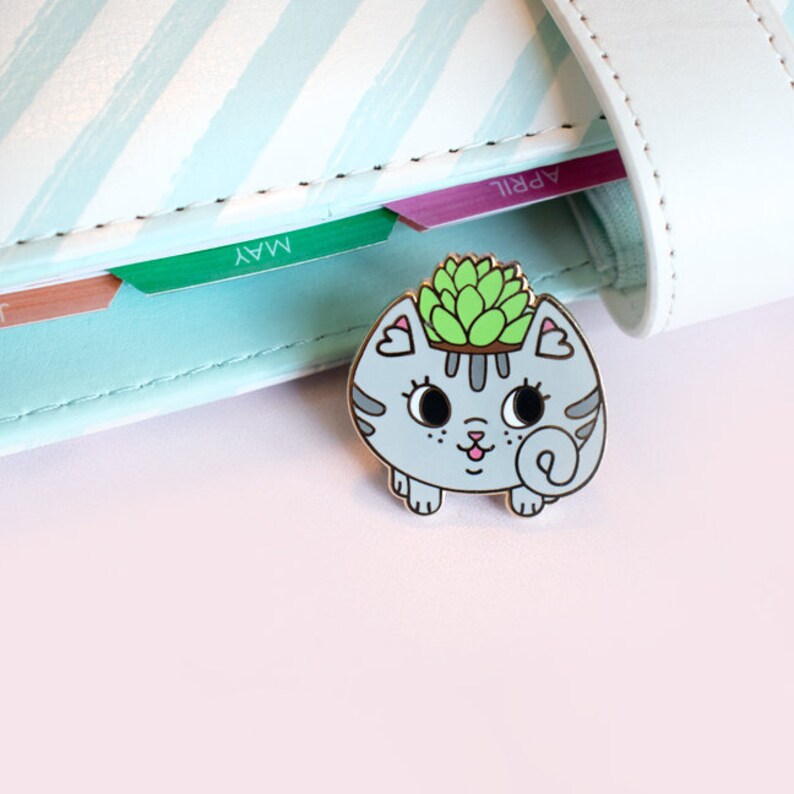 May include: A grey and white enamel pin of a cat with a succulent plant growing out of its head. The cat is smiling and has a pink nose. The pin is on a light blue and white striped background.