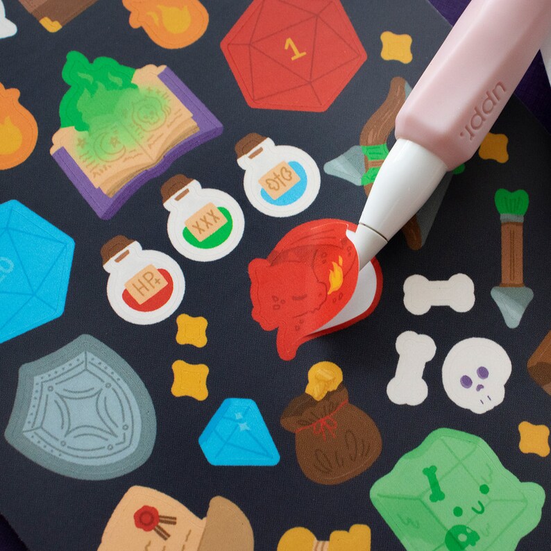 May include: A sheet of stickers with a variety of fantasy and role-playing game themed designs. The stickers include a red dragon, a blue gem, a green potion bottle, a gold coin, a shield, a dice, and a book. The stickers are on a black background.