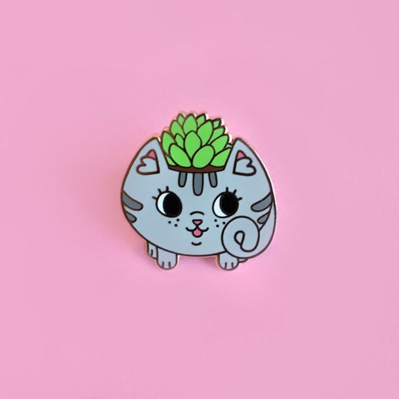 May include: A grey cat planter with a green succulent plant growing out of the top. The cat planter has a cute cartoon face with black eyes and a pink nose.