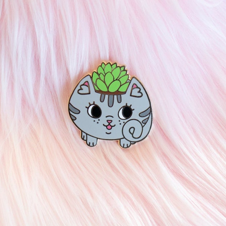 May include: A grey cat-shaped planter with a green succulent plant growing out of the top. The planter has a cute cartoon face with pink cheeks and a curled tail.