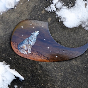 Könnte beinhalten: Ein Gitarrenplektrum aus Holz mit einem gemalten Bild eines Wolfes, der zum Mond heult, mit Sternen im Hintergrund.