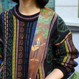 May include: A brown leather guitar strap with a green leaf pattern and a cartoon illustration of a person playing a guitar.