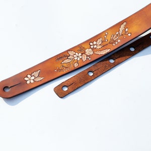 May include: A brown leather guitar strap with a floral design. The strap has multiple holes for adjusting the length.