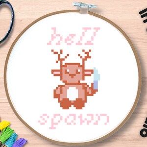 Puede incluir: Un aro de bordado de punto de cruz con una imagen pixelada rosa y marrón de un ciervo sosteniendo un cuchillo. El texto "hell spawn" está bordado en rosa debajo del ciervo. El texto "In Stitches Studio" está escrito verticalmente en la esquina superior derecha de la imagen.