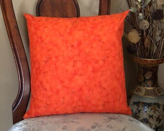 May include: Orange throw pillow with a subtle pattern of small white dots. The pillow is sitting on a chair with a floral patterned fabric.