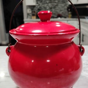 May include: A vibrant red ceramic pot with a matching lid and a metal handle. The pot has a rounded body, three small feet, and a glossy finish. This decorative piece is suitable for kitchen or home decor.