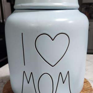 Puede incluir: Un tarro de cerámica azul claro con tapa, con la frase "I ❤️ MOM" en letras negras. El tarro está sobre un soporte de madera con borde de cuentas y una cinta crema atada al asa.