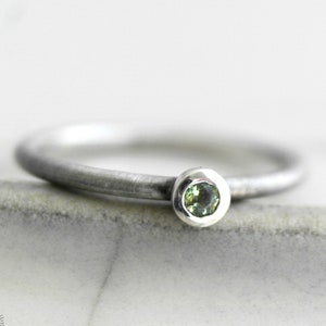 Può includere: Un semplice anello a fascia d'argento con una piccola pietra preziosa verde rotonda incastonata in una montatura a castone.