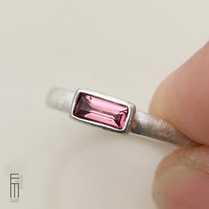 May include: A silver ring featuring a rectangular, pink gemstone. The gemstone is set in a rectangular bezel. The ring has a brushed metal finish and is held by a person's fingers.