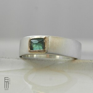 May include: A silver band ring with a square green gemstone set in a gold bezel. The ring has a brushed finish.