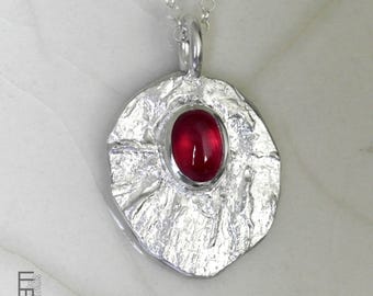 Op de afbeelding: Zilveren hangerketting met een getextureerde, onregelmatig gevormde hanger met een heldere rode ovale edelsteen in het midden. De hanger hangt aan een fijne zilveren ketting, waardoor een opvallend contrast ontstaat.
