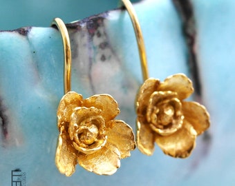 Può includere: Un paio di orecchini color oro con un design floreale dettagliato, petali testurizzati e un bocciolo centrale. Ogni orecchino è fissato a un gancio curvo. Il logo di Ellen Moiffer è visibile nell'angolo in basso a sinistra dell'immagine.