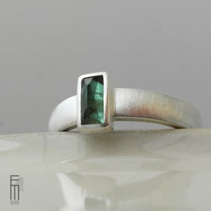 May include: A silver ring with a green gemstone set in a rectangular bezel. The ring has a brushed finish.