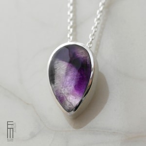 Könnte beinhalten: Eine silberne Halskette mit Anhänger, die einen tropfenförmigen Edelstein in Lila- und Weißtönen zeigt. Der Edelstein ist in eine polierte Silberfassung gefasst und hängt an einer zarten Silberkette. Die Halskette wird auf einer weißen Marmoroberfläche präsentiert.