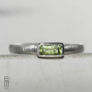 May include: A silver ring featuring a rectangular, pale green gemstone. The ring has a brushed metal finish and a simple design. The gemstone is set in a bezel setting, and the band is thin and smooth.