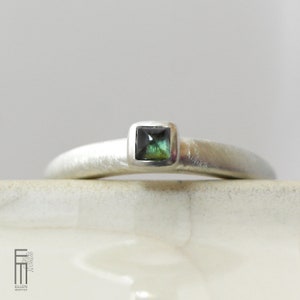 May include: A silver ring with a square, dark green gemstone. The ring has a brushed metal finish and a simple, elegant design. The gemstone is set in a bezel setting, and the band is smooth and rounded.