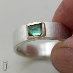 May include: A silver band ring with a green gemstone set in a gold bezel. The ring has a brushed finish.