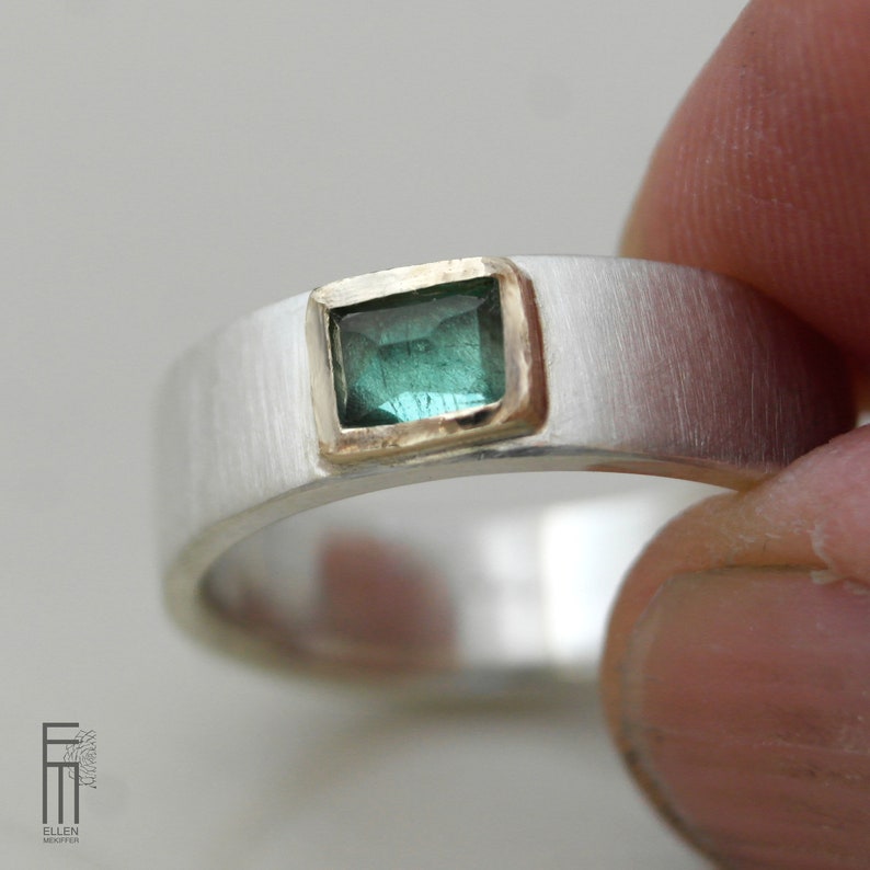 May include: A silver band ring with a green gemstone set in a gold bezel. The ring has a brushed finish.