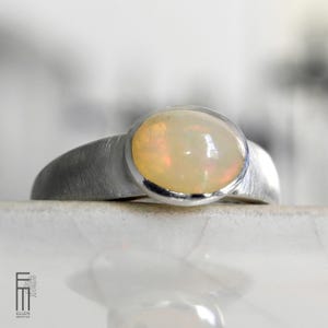 Könnte beinhalten: Ein silberner Ring mit einem großen, ovalen Opal-Edelstein. Der Opal hat ein cremiges, schillerndes Aussehen, gefasst in einem glatten, gebürsteten Silberband. Der Ring wird auf einer weißen Oberfläche präsentiert, die das Design des Schmucks hervorhebt.