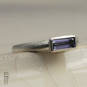 May include: A silver ring with a rectangular, deep purple gemstone. The ring has a brushed metal finish and a simple, elegant design. The gemstone is set in a bezel setting, and the band is smooth and rounded.