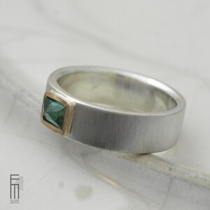 May include: A silver band ring with a green gemstone set in a gold bezel. The ring has a brushed finish.