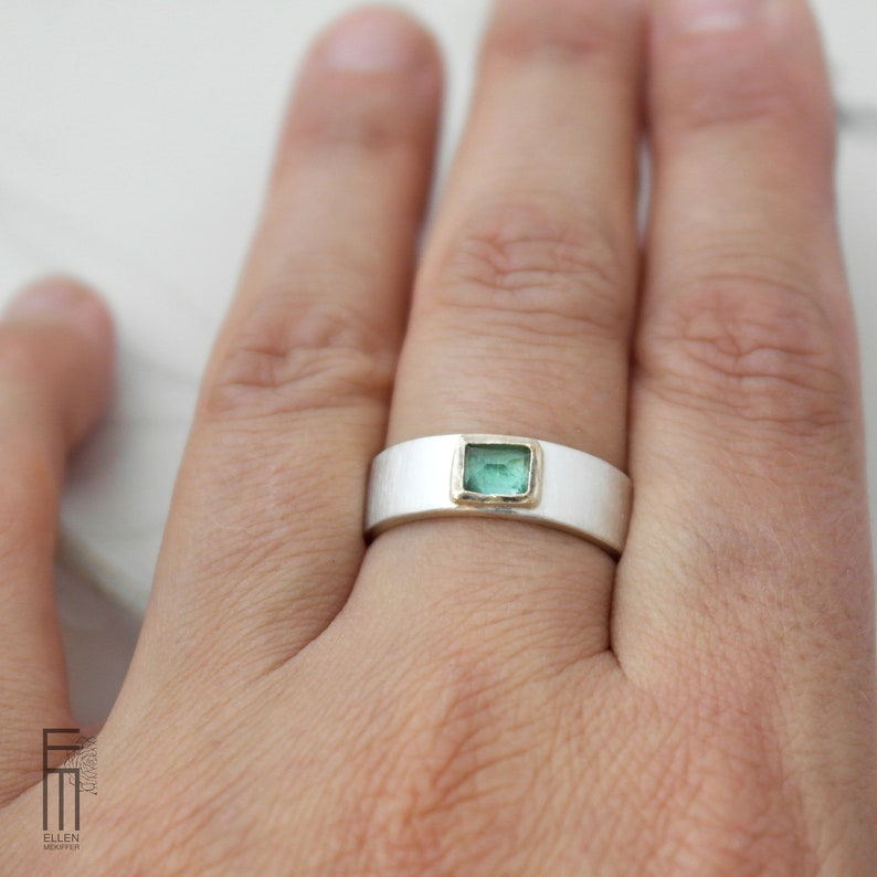 May include: A silver band ring with a square green gemstone set in a gold bezel. The ring is worn on a person's finger.
