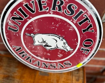 May include: Round metal sign with the words "UNIVERSITY OF ARKANSAS" in black lettering on a red background. A white Razorback logo is in the center. The sign has a distressed, vintage look.