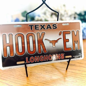 May include: A rectangular license plate with the word "TEXAS" at the top and "HOOK 'EM LONGHORNS" in large, bold letters. The plate features a burnt orange and white color scheme, with the Longhorns logo prominently displayed. It is held by a black metal stand.