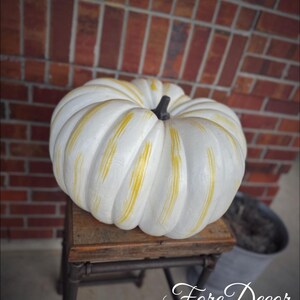 May include: A large, white pumpkin with yellow accents and a dark stem sits on a small, rustic wooden table. The pumpkin is the focal point, with a brick wall in the background. The text "Fore Decor" is in the lower right corner.