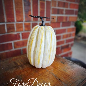 May include: A decorative white pumpkin with vertical yellow stripes and a black stem. The pumpkin is sitting on a wooden table. The word "Fore Decor" is written on the table.