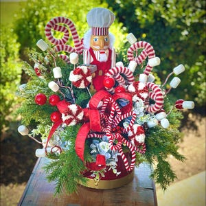 May include: A Christmas centerpiece featuring a nutcracker figurine, red and white candy canes, white marshmallows, and red berries in a red and gold pot.
