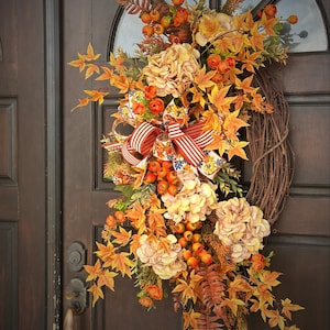 May include: A decorative wreath featuring a grapevine base adorned with artificial autumn foliage, including orange maple leaves, cream-colored hydrangea blossoms, and clusters of orange berries, accented by a striped red and white fabric bow.