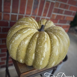 May include: A large, decorative pumpkin in shades of olive green with white speckles and vertical stripes. The pumpkin has a dark stem and sits on a small, rustic wooden table. The text "Fore Decor" is visible in the image.
