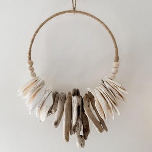 May include: A circular wall hanging with a natural design. The ring is wrapped in twine and decorated with wooden beads, white shells, and driftwood pieces. The shells and driftwood are arranged in a decorative pattern.
