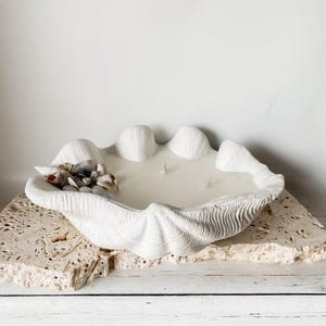 May include: A large, white clam shell-shaped candle with three wicks. The candle is filled with a creamy wax and decorated with small seashells. The candle sits on a textured, light-coloured stone base.