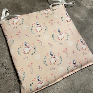 May include: Square seat cushion with a light pink background featuring a repeating pattern of ducks wearing hats and floral wreaths. White ribbon ties are attached to each corner. The cushion is approximately 16 inches square.