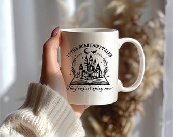 Può includere: Tazza in ceramica bianca con un disegno nero di un castello all'interno di un libro aperto. Il testo recita "I still read fairy tales they're just spicy now".