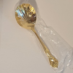 Puede incluir: Una cuchara de servir dorada con un cuenco decorativo con forma de concha y un mango detallado. La cuchara está empaquetada en una bolsa de plástico transparente.