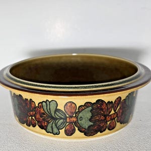 Peut inclure: Un bol rond en céramique avec un motif floral. Le bol a une base jaune clair avec un motif répété de fleurs marron, vertes et marron. Le bord est marron avec une bande verte.