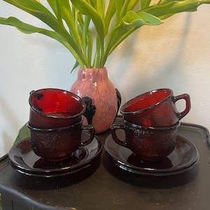 Può includere: Due set di tazze da tè e piattini in vetro rosso. Ogni set ha tre tazze impilate l'una sull'altra. Le tazze hanno una superficie testurizzata e sono di un rosso intenso. I piattini sono di un rosso abbinato.