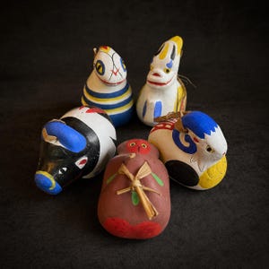 May include: A collection of five hand-painted, folk art figurines. The figurines are in the shape of animals and other objects, with bright colors and detailed designs. The figures are arranged on a dark background.