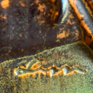 K&ouml;nnte beinhalten: Nahaufnahme eines Keramikst&uuml;cks mit strukturierter Oberfl&auml;che. Das St&uuml;ck weist eine Mischung aus Dunkelbraun, Orange und Gr&uuml;nt&ouml;nen auf. Das Wort "Lena" ist in einer stilisierten Schriftart eingraviert.