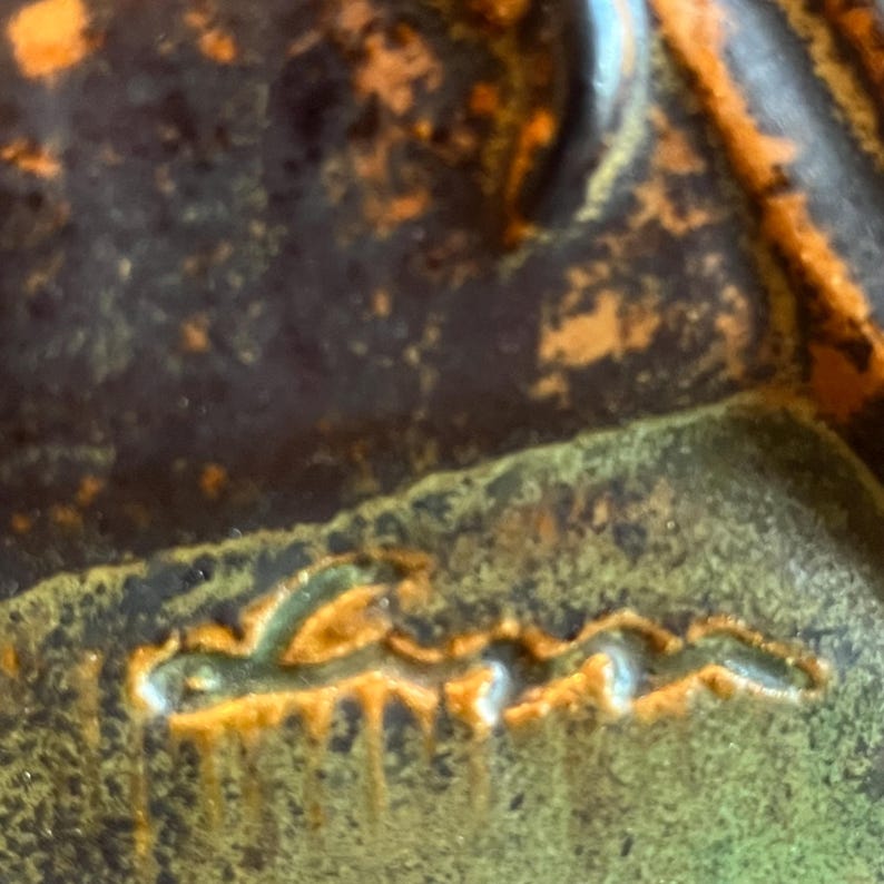 K&ouml;nnte beinhalten: Nahaufnahme eines Keramikst&uuml;cks mit strukturierter Oberfl&auml;che. Das St&uuml;ck weist eine Mischung aus Dunkelbraun, Orange und Gr&uuml;nt&ouml;nen auf. Das Wort "Lena" ist in einer stilisierten Schriftart eingraviert.