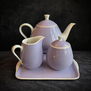 Puede incluir: Un juego de té de cerámica lavanda con tetera, crema, azucarero y bandeja. La tetera tiene un asa y un pico blancos. La crema y el azucarero tienen detalles blancos. El juego está sobre una bandeja lavanda con ribete blanco.