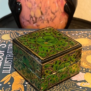 May include: A small, square, wooden box with a hinged lid. The box is painted black with green floral designs and gold accents. The box is sitting on a book with a dark blue cover.