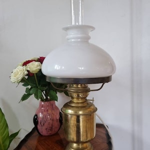 Może przedstawiać: Antyczna lampa naftowa wykonana z mosiądzu z białym szklanym kloszem. Lampa ma ozdobną mosiężną podstawę i szklany komin. Lampa stoi na drewnianym stole.