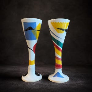 Puede incluir: Dos copas de vino de cerámica blanca pintadas a mano con diseños geométricos coloridos. Una copa presenta formas azules, amarillas y negras, mientras que la otra tiene rayas amarillas, verdes, rojas y azules. Las copas tienen una parte superior acampanada y un tallo delgado.