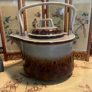 Peut inclure: Une théière en céramique avec un couvercle et une anse. La théière est principalement marron et beige, avec un motif marron plus foncé sur le couvercle. L'anse est blanche. La théière est probablement vintage ou antique.