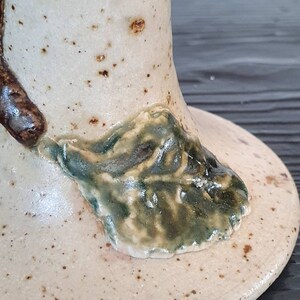 May include: A ceramic vase with a speckled white glaze and a green leaf design. The vase has a wide base and a narrow neck.